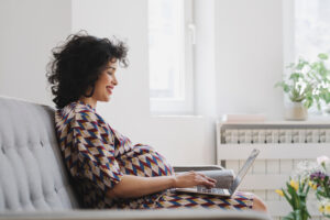 Beautiful pregnant woman sitting on couch at home and typing on her laptop.